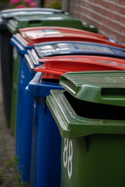 An outdoor storage area filled with stacks of flattened cardboard boxes displaying branded fruit packaging, placed on wooden pallets, and surrounded by large green and red waste bins with closed lids.